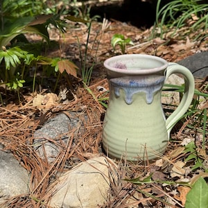 Puede incluir: Una taza de cerámica con cuerpo verde claro moteado y borde acampanado con un patrón de esmalte de goteo azul y morado. La taza tiene un asa curva en el lado derecho y una base redondeada, mostrando un diseño de alfarería artesanal.