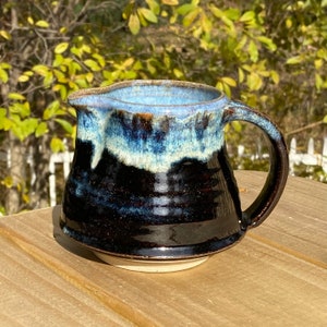 May include: A dark brown ceramic creamer with a blue and white glaze. The creamer has a rounded body and a handle.