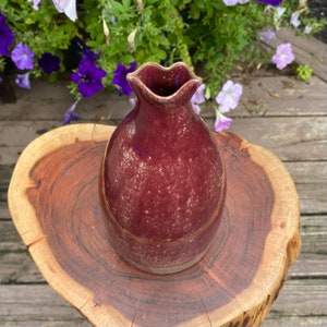 May include: A small, burgundy ceramic vase with a speckled glaze. The vase has a unique, wavy rim and a rounded body.