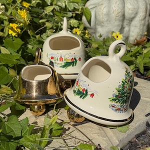 May include: Three ceramic bell-shaped ornaments. One is gold, the others are white with a gold trim and feature a winter village scene with a Christmas tree. The bells have a loop at the top and a round base.