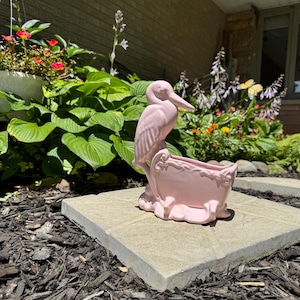 May include: A light pink ceramic planter shaped like a bird standing next to a basin. The bird has a long neck and pointed beak, while the basin features decorative scrollwork along the rim and rests on a base with small, rounded feet.
