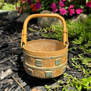 May include: A handmade ceramic basket with a bamboo handle. The basket is tan with a band of blue-green square designs around the middle. The handle is attached with leather-like material. The basket is sitting on black mulch.