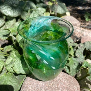 Puede incluir: Un pequeño jarrón de vidrio verde con remolinos y motas blancas. El jarrón tiene un fondo redondeado y una abertura ancha y acampanada.