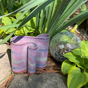 May include: A ceramic vase with a unique, undulating, ribbon-like shape. The surface features a glossy glaze in blended shades of lavender, soft blue, and pale green, with a visible terracotta-colored base rim and a smooth, curved opening.