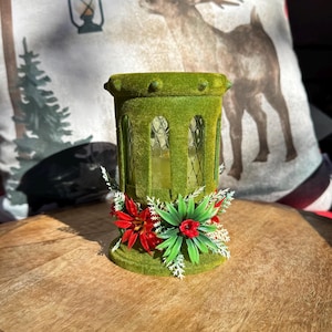 May include: A green, vintage-style candle holder with a textured, velvety surface. The candle holder has arched windows and is decorated with red and green floral accents. The overall design evokes a classic, festive aesthetic.