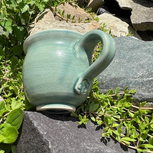 Op de afbeelding: Een turquoise keramische mok met een craquelé-afwerking. De mok heeft een grote handgreep met een bloemmotief. De mok staat op een donkergrijze rots, omringd door groen.