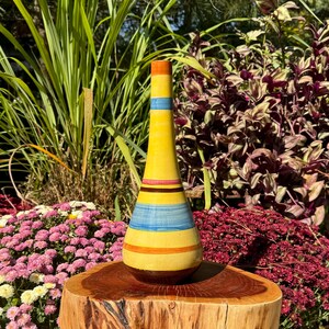 May include: A yellow ceramic vase with blue, orange, and brown stripes. The vase is sitting on a wooden stump.