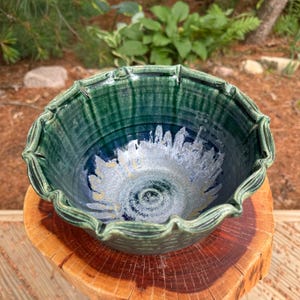 May include: A handcrafted ceramic bowl with a scalloped rim. The bowl showcases a colour gradient, shifting from a deep teal green at the edge to a dark blue centre with a silver and white floral pattern. The bowl is displayed on a wooden surface.