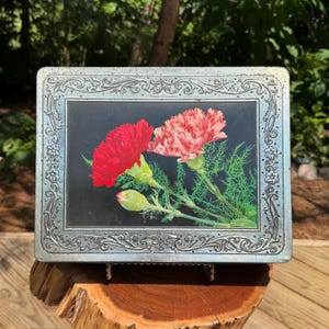 May include: Rectangular metal tin with a floral design. The center features a black background with red and pink carnations and green foliage. The tin has a decorative silver frame with ornate detailing. The tin is displayed on a wooden surface.