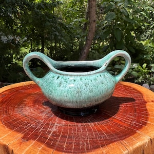 Peut inclure: Un bol en céramique vintage avec deux anses incurvées, émaillé en turquoise moucheté et marron foncé. Le bol repose sur une base sombre et est présenté sur une surface en bois, mettant en valeur son design unique.