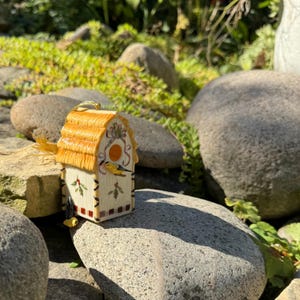 May include: A decorative birdhouse with a straw roof and a white body. The birdhouse is adorned with painted birds, holly leaves, and a circular design. The birdhouse is sitting on a rock in a garden setting.