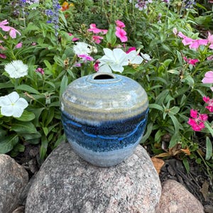 May include: A round ceramic vase with a blue, gray, and tan color scheme. The vase has a dark blue band with a lighter blue stitched design. The vase is sitting on a large gray rock, surrounded by colorful flowers.