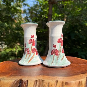 May include: Two white ceramic candle holders with a floral design. The candle holders have a flared base and a narrow neck. The floral design features red flowers with brown stems and green leaves. The rims are trimmed with a green speckled pattern.