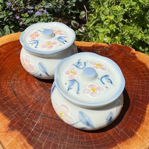 Op de afbeelding: Twee witte keramische containers met blauwe en roze bloemenmotieven. De containers hebben deksels met kleine gaatjes en zijn perfect om kleine spullen in op te bergen.