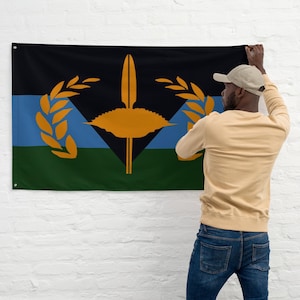 May include: A rectangular flag with a black top, blue middle, and green bottom. The flag features a central design of a yellow spear-like shape, a diamond shape, and two yellow laurel branches. The flag is being held up against a white wall.