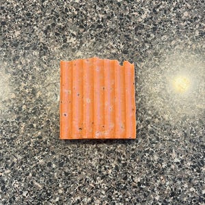 Puede incluir: Una barra de jabón rectangular de color naranja con una superficie texturizada. El jabón tiene un tono rosa claro y está salpicado de pequeños puntos blancos.
