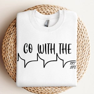 May include: White sweatshirt with the text "GO WITH THE" above a speech bubble design, and "RRT LIFE" in a smaller font. The sweatshirt is displayed on a woven, circular surface.
