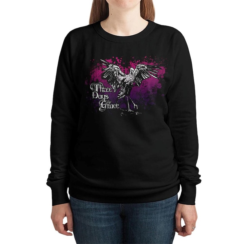 May include: Black sweatshirt featuring a white and gray illustration of a bird with outstretched wings, set against a purple and pink splatter design. The text "Three Days Grace" is printed in white, in a stylized font.