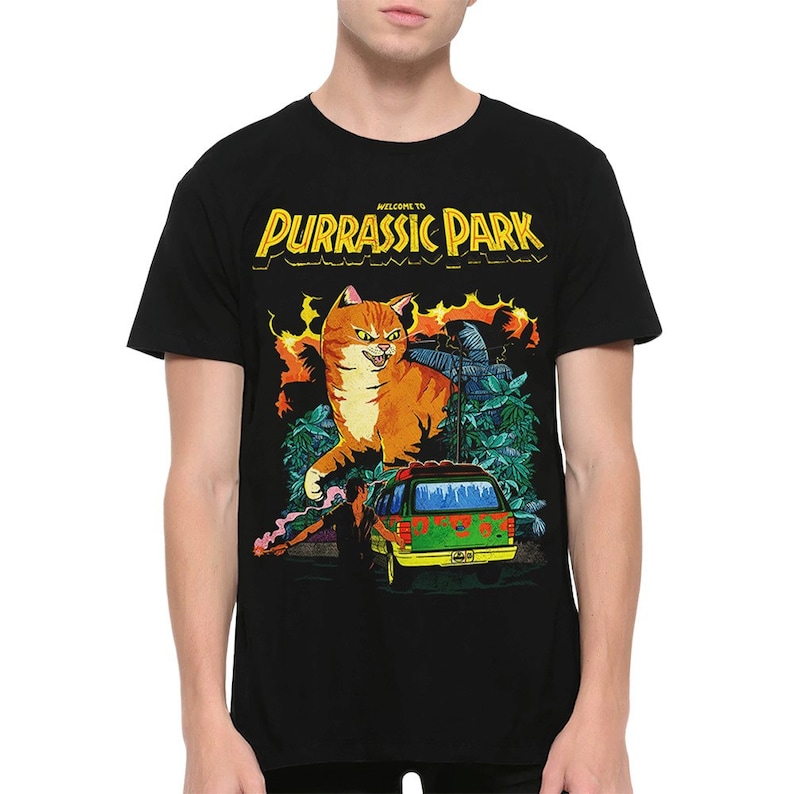K&ouml;nnte beinhalten: Schwarzes T-Shirt mit einem Grafikdruck eines gro&szlig;en orangefarbenen Katers mit gr&uuml;nen Augen, einem gr&uuml;nen Van und dem Text "Welcome to Purrassic Park".