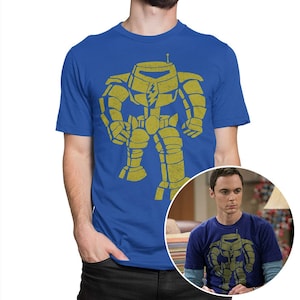 Op de afbeelding: Een koninklijk blauw t-shirt met een vintage-stijl afbeelding van een gele robot. De robot heeft een bliksemflits op zijn borst. Het t-shirt wordt getoond op een model dat een zwarte spijkerbroek draagt.