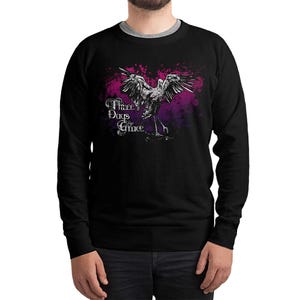 May include: Black crew neck sweatshirt featuring a graphic of a bird with outstretched wings, set against a purple and pink splatter design. The text "Three Days Grace" is printed in white script. A casual, comfortable apparel item.