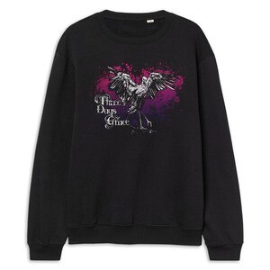 May include: Black sweatshirt featuring a graphic of a bird with outstretched wings, set against a pink and purple background. The text "Three Days Grace" is displayed in a stylized font. A crew neck sweatshirt.