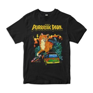 K&ouml;nnte beinhalten: Schwarzes T-Shirt mit einem Grafikdruck eines gro&szlig;en orangefarbenen Katers mit gelben Augen und einem bedrohlichen Ausdruck. Die Katze steht vor einem gr&uuml;nen Van mit dem Text "Willkommen im Purrassic Park" dar&uuml;ber.