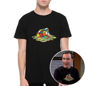 Op de afbeelding: Een zwart T-shirt met een kleurrijke, smeltende Rubik's Cube-afbeelding. De afbeelding toont de kubus die smelt in een plas kleurrijke vloeistof.