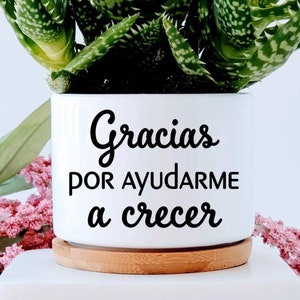 May include: White ceramic planter with a wood base. The planter has the text "Gracias por ayudarme a crecer" on the front and a red heart and the name "Jessie" on the back. The planter is filled with a green succulent plant.