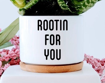 May include: White ceramic planter with a wooden base. The planter has black text that reads "Rootin for you". A green succulent plant is growing in the planter.