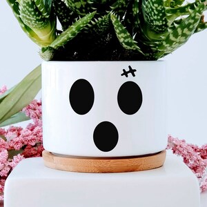 May include: White ceramic planter with a ghost face design. The planter has three black circles for eyes and a mouth, and a small black cross for a nose. The planter is filled with a green succulent plant.