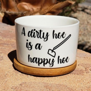 May include: White ceramic planter with a wooden base. The planter has a black graphic that reads "A dirty hoe is a happy hoe" with a small image of a garden hoe.