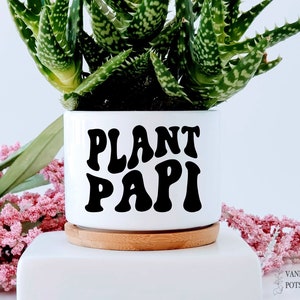 May include: White ceramic planter with a black "PLANT PAPI" graphic in a retro font. The planter has a wooden base and is filled with a green succulent plant.