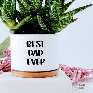 May include: White ceramic planter with a wood base. The planter has black text that reads "Best Dad Ever". A green succulent plant is growing in the planter.