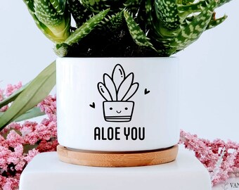 May include: White ceramic planter with a black and white illustration of a succulent with the text "Aloe You". The planter has a wooden base and is filled with a green succulent plant.