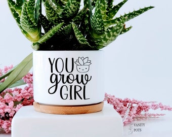 May include: White ceramic planter with a wood base, featuring a green succulent plant and the text "YOU grow GIRL" in black lettering.