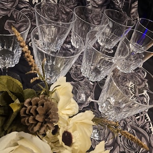 May include: A selection of clear, patterned drinking glasses on a black and white floral patterned surface. The glasses are complemented by white flowers, a pinecone, and dried plant sprigs.