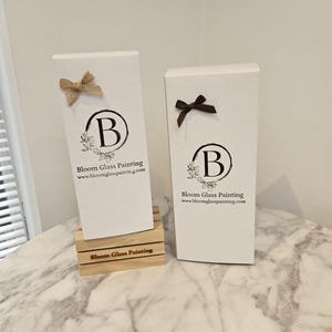 May include: Two white boxes with the text "Bloom Glass Painting" and the website address "www.bloomglasspainting.com". The boxes feature a brown ribbon and the letter "B" with a floral design. The box on the left is on a wooden base with the text "Bloom Glass Painting".