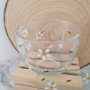 May include: A glass mug and coaster set featuring a daisy and bee design. The mug and coaster are both clear glass with white daisies and yellow bees painted on them. The daisies have green centres and the bees have black stripes.