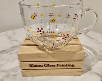 May include: A clear glass mug with a hand-painted design of bees and flowers. The mug is sitting on a wooden coaster with the words "Bloom Glass Painting" engraved on it.