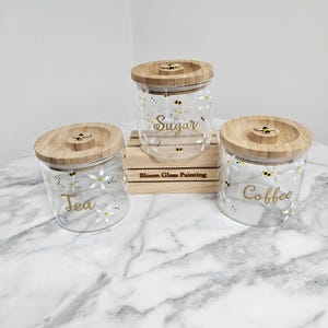 May include: Three glass canisters with wooden lids and bee and daisy designs. The canisters are labelled "Tea", "Sugar", and "Coffee".