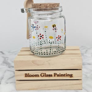 May include: A clear glass jar with a cork lid, decorated with hand-painted flowers and a bee motif. A small wooden spoon is attached. The jar is on a wooden crate with the text "Bloom Glass Painting".