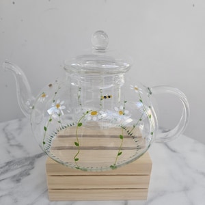 May include: A clear glass teapot featuring a hand-painted design of white daisy flowers with yellow centres, green stems, and small bees. The teapot includes a matching glass lid and an internal glass infuser basket for loose leaf tea brewing.