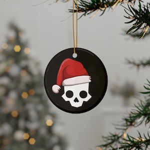 May include: A black, round Christmas ornament featuring a white skull wearing a red Santa hat with a white trim. The ornament hangs from a gold string, with a blurred Christmas tree in the background.