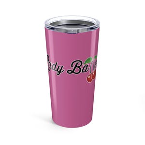 Puede incluir: Un vaso de viaje rosa con tapa plateada. El vaso presenta el texto "Lady Baller" en una fuente cursiva con un gráfico de cereza. El vaso está diseñado para mantener las bebidas calientes o frías.