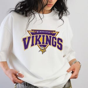 Könnte beinhalten: Ein weißes kurzärmeliges T-Shirt mit einer violetten und goldenen Grafik auf der Brust. Die Grafik zeigt den Text Minnesota Vikings in einer fetten, geschwungenen Schrift über einem dreieckigen Emblem mit einem Wikingerhelm-Symbol.