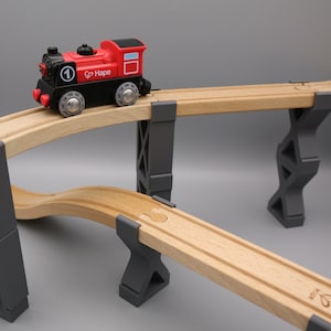 Op de afbeelding: Een rode en zwarte speelgoedtrein met het woord "Hape" aan de zijkant, die over een houten treinspoor rijdt met grijze houten steunen.