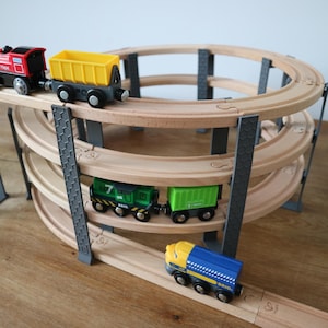 Puede incluir: Juego de tren de juguete de madera con una pista de varios niveles. El juego incluye un tren rojo, un vagón amarillo, un tren verde, un vagón verde y un tren azul y amarillo. La pista es de madera marrón claro y está sostenida por pilares grises.