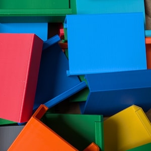 Könnte beinhalten: Eine Ansammlung farbenfroher, geometrischer 3D-gedruckter Objekte in Blau-, Grün-, Rot-, Orange- und Gelbtönen. Die Objekte haben eine rechteckige Form mit einer Lippe, was auf Tabletts oder Behälter hindeutet.