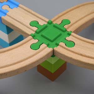 Könnte beinhalten: Ein hölzernes Spielzeug-Eisenbahnset mit einem grünen, vierseitigen Kreuzungsteil. Die Schienen sind aus Naturholz und das Kreuzungsteil aus Kunststoff.